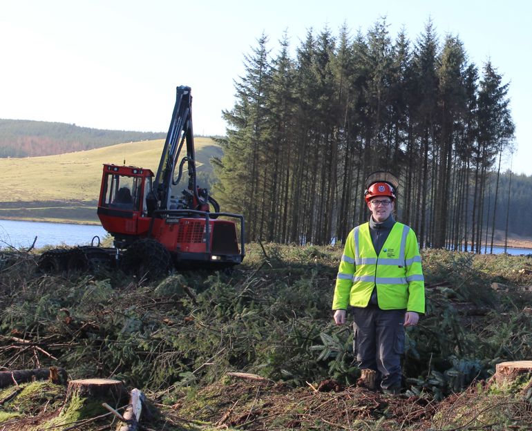Man in a high visibility jacket stands in front of a digger and a small group of trees
